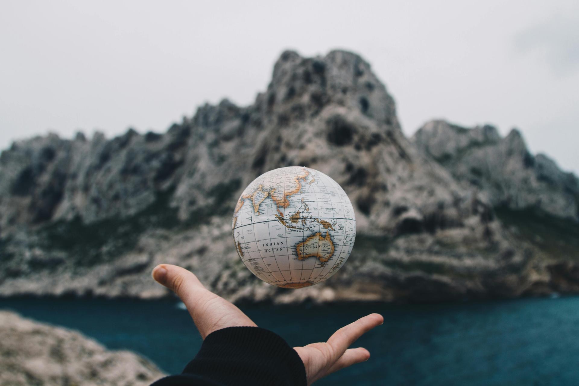 A hand holds a small globe against a coastal landscape